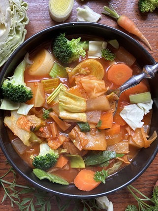Auf einem Holztisch steht eine schwarze Schale mit Kohlsuppe. Um die Schale herum liegen Brokolli, Karotte, Zwiebel und Kohl.