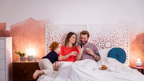 Kind sitzt bei Eltern mit Smartphone im Bett. Smartphones Kinder Gefahren