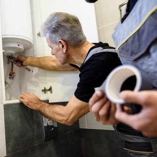 Grauhaariger Mann schraubt an Boiler rum, daneben steht ein jüngerer Mann in Arbeitshosen mit Klebeband in der Hand. Unseriöse Portale vermitteln Handwerker, die schlecht arbeiten und total überzogene Rechnungen stellen.