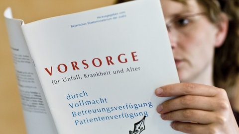 Unscharf im Hintergrund zu sehende, mittelalte Person mit Brille liest Broschüre über Vorsorge. Neues Betreuungsrecht 2023: Patientenverfügung, Vorsorgevollmacht, Betreuungsverfügung, gesetzlicher Betreuer