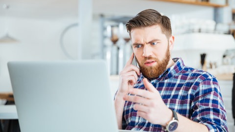 Mann hält in der rechten Hand ein Handy. Er sitzt vor einem Laptop und blickt grimmig hinein.