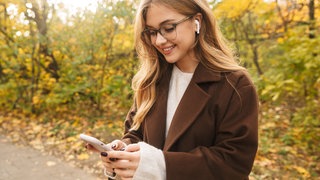 Frau mit blonden, langen Haaren und einer Brille schaut auf ihr Smartphone trägt dabei Airpods
