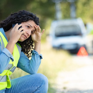 Eine Frau sitzt in Warnweste vor ihrem Auto und ruft jemanden an. Durch reflektierende Farben sollen Warnwesten in Notsituationen Sicherheit geben. Doch was taugt das leuchtende Material, wenn es darauf ankommt?   