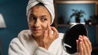 Eine Frau hält sich einen Handspiegel vors Gesicht und prüft die Haut auf mögliche Falten.