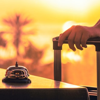Frau hält den Griff ihres Koffers, Rezeptionsklingel, Urlaubsszenerie im Hintergrund
