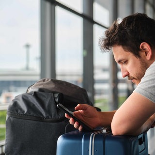 Mann sitzt mit Koffern am Flughafen und schaut auf sein Handy