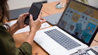 Eine Frau sitzt vor einem Laptop mit einem Online-Shop und tippt auf ihrem Smartphone.