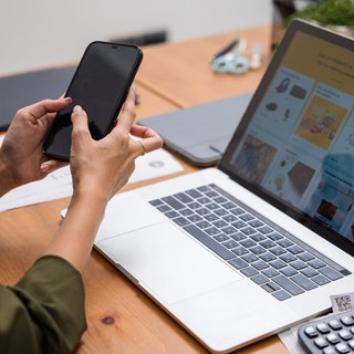 Eine Frau sitzt vor einem Laptop mit einem Online-Shop und tippt auf ihrem Smartphone.