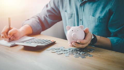 Mann sitzt am Schreibtisch mit Sparschwein, Taschenrechner, Geld und Notizblock