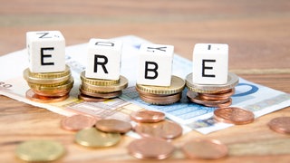 Mit Buchstaben-Würfeln wird das Wort "Erbe" gelegt. Die Würfel liegen auf Geldscheinen und Münzen.