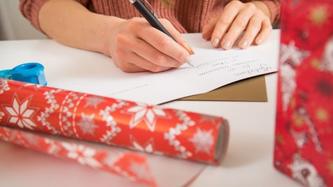 Eine Frau schreibt eine Weihnachtskarte | Weihnachten 2025: Das sind die Versandfristen für Pakete und Briefe