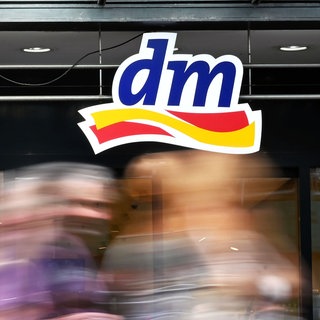 Logo der Drogeriemarktkette DM, im Vordergrund laufen Menschen vorbei. Die Drogerie startet ins Apothekengeschäft und verkauft nun auch verschreibungsfreie Medikamente.