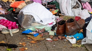 Altkleider neben einem Altkleider-Container mit verschiedenen Kleidungsstücken wie braunen Lederschuhen, Sandalen oder Hosen zeigen die Wegwerfgesellschaft und Vergänglichkeit von Mode und Kleidung