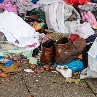 Altkleider neben einem Altkleider-Container mit verschiedenen Kleidungsstücken wie braunen Lederschuhen, Sandalen oder Hosen zeigen die Wegwerfgesellschaft und Vergänglichkeit von Mode und Kleidung