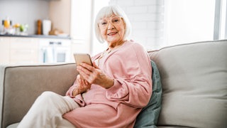 Eine alte Frau sitzt lächelnd auf einer Couch und hält ein Smartphone in ihrer Hand.