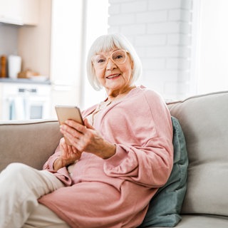 Eine alte Frau sitzt lächelnd auf einer Couch und hält ein Smartphone in ihrer Hand.