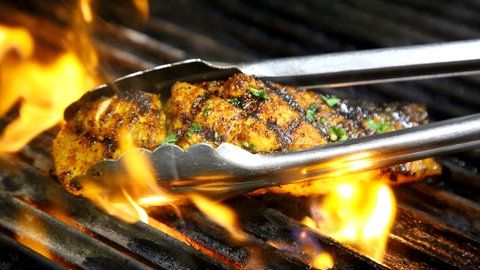 Ein Stück Fisch liegt auf einem Holzkohlegrill und wird mit einer Grillzange gewendet.