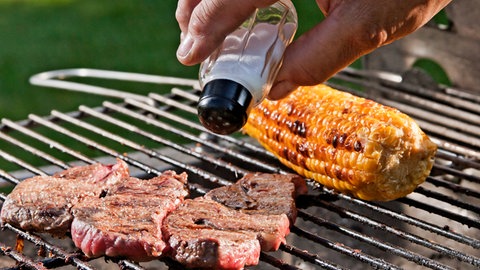 Mehrere Steaks liegen auf einem Grill und werden mit Salz bestreut. Außerdem liegt ein Maiskolben auf dem Grill.