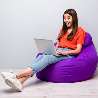 Junge Frau auf lilafarbenem Sitzsack schaut mit freudig-überraschtem Gesicht in Laptop auf ihrem Schoß. Live-Shopping auf Tic Toc oder Instagram wird immer beliebter.