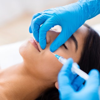 Weibliches Gesicht, in das behandschuhte Hände eine Spritze injizieren. Welche Risiken gibt es bei Hyaluron?