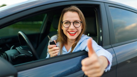 Junge Frau schaut aus Autofenster und hält stolz Autoschlüssel vor sich. Tipps und Tricks für den Gebrauchtwagenkauf. 