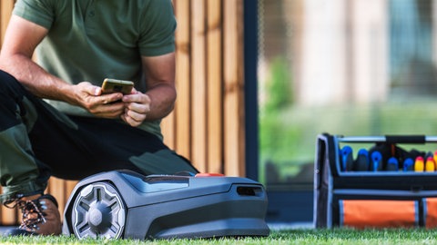 Mann kniet neben Mähroboter und hält Smartphone in der Hand.