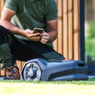 Mann kniet neben Mähroboter und hält Smartphone in der Hand.