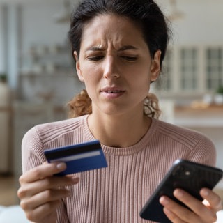 Frau mit verwirrtem Gesichtsausdruck, in einer Hand Handy, in der anderen Hand Kreditkarte.