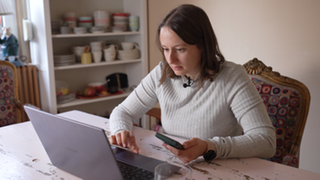 Naomi sitzt an ihrem Laptop mit dem Handy in der Hand 