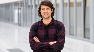 Ein Mann mit langen Haaren steht mit verschränkten Armen und in legerem Hemd in einer hellen Halle und lächelt