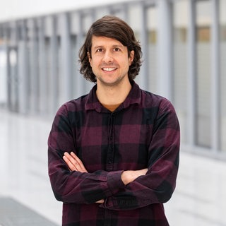 Ein Mann mit langen Haaren steht mit verschränkten Armen und in legerem Hemd in einer hellen Halle und lächelt