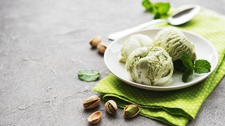 Drei Kugeln Pitazieneis auf einem kleinen Teller. Auf dem Tisch liegen eine Serviette, ein kleiner Löffel, Pistazien und Minzblätter.
