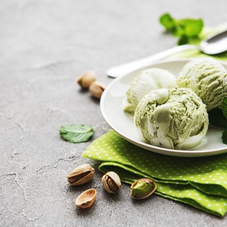 Drei Kugeln Pitazieneis auf einem kleinen Teller. Auf dem Tisch liegen eine Serviette, ein kleiner Löffel, Pistazien und Minzblätter.
