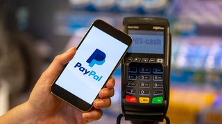 Handy mit Paypal-App an einer Kasse: Der in Deutschland beliebte amerikanische Zahlungsdienstleiter Paypal bietet jetzt auch kontaktloses Bezahlen an -  eine Art virtuelle Kreditkarten auf dem Smartphone.