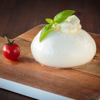 Burrata auf einem Schneidebrett garniert mit Basilikum neben einer Cocktail-Tomate