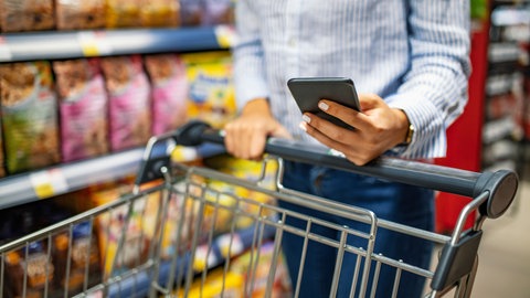 Auf dem Bild sieht man den Oberkörper einer Person, die beim einkaufen einen Einkaufswagen schiebt und ihr Handy in der Hand hält. Punkte sammeln, Schnäppchen machen - mit Supermarkt-Apps ist das möglich. Doch Vorsicht vor Betrügern, die Bonuspunkte klauen! So können sich Verbraucher schützen! 