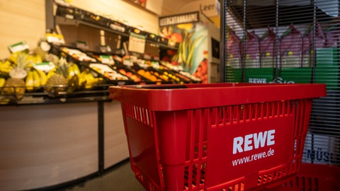 Auf dem Bild ist ein Rewe-Korb zu sehen, im Hintergrund sind Supermarkt-Regale. Punkte sammeln, Schnäppchen machen - mit Supermarkt-Apps ist das möglich. Doch Vorsicht vor Betrügern, die Bonuspunkte klauen! So können sich Verbraucher schützen! 