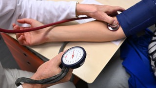 Ein Arzt misst in einem Untersuchungsraum bei einer Patientin den Blutdruck.Die gesetzlichen Krankenversicherungen werden immer teurer. Ab Juli erhöhen sechs weitere Krankenkassen ihre Zusatzbeiträge.