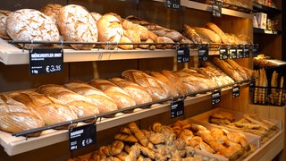 Brot im Regal einer Bäckerei. Brot und Brötchen – kaum ein Lebensmittel ist uns so lieb wie das tägliche Gebäck aus der Bäckerei. Doch was steckt tatsächlich hinter dem, was uns als Handwerk verkauft wird?