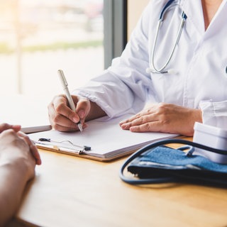 Arzt in weißem Kittel sitzt an Schreibtisch gegenüber Patient, nur Oberkörper sichtbar. Das "Geschäft" mit der Dringlichkeit bei Facharztterminen.