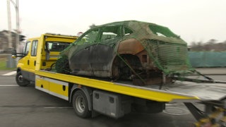 Abgebranntes Autowrack eines Mercedes EQA ist mit Gurten und einen Netz auf einem gelben Abschleppwagen fixiert.