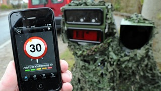 Ein Smartphone mit einer Blitzer-Warn-App wird vor eine mobile Geschwindigkeits-Messanlage gehalten. 