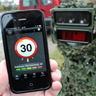 Ein Smartphone mit einer Blitzer-Warn-App wird vor eine mobile Geschwindigkeits-Messanlage gehalten. 