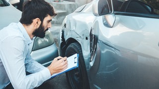 Mann mit Klemmbrett kniet vor Auto mit Macken. Kürzen Kfz-Haftpflichtversicherungen häufig Leistungen?