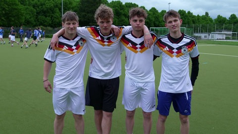 Vier junge Männer mit Deutschlandtrikots stehen Arm in Arm auf Fußballplatz. Fußball-Trikots für die WM 2026 - Original versus Fakes