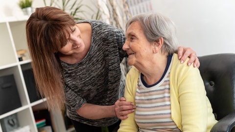 Mittelalte Frau bäugt sich freundlich über alte Frau. Wie die Pflegekasse24 mit Online-Pflegekursen Kasse macht.