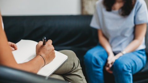 Coaching: Zu sehen sind zwei Personen ohne ihr Gesicht. Im Vordergrund eine Person mit Stift und Papier, im Hintergrund, in der Unschärfe, eine Person die auf einem schwarzen Sofa sitzt. 