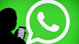 Ein Mann mit Smartphone steht vor einem Monitor mit dem Logo des Messenger-Dienstes WhatsApp.