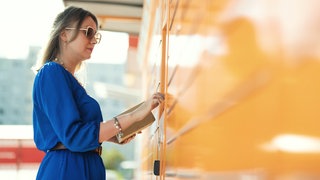 Frau an Packstation: In Packstationen kann es im Sommer um die 60 Grad heiß werden – schlecht für Schokolade, Kosmetik oder Elektronik. Was das für Ihre Rechte bedeutet, und wie Sie ohne App an Ihr Paket kommen.