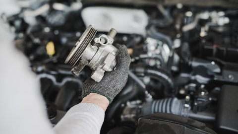 Zu sehen ist die Hand einer Person, die vor einem Auto mit geöffneter Motorhaube steht und ein Autoersatzteil hält.